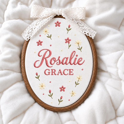 May include: An oval wooden embroidery hoop containing white fabric with the embroidered name Rosalie GRACE in pink script and block lettering. The design features small pink and yellow flowers with green stems and a white lace bow at the top.