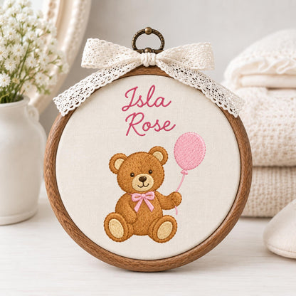 May include: A circular wooden embroidery hoop containing white fabric with the embroidered name Isla Rose in pink script. Below the text is a brown teddy bear with a pink bow tie holding a pink balloon. A cream lace bow is tied at the top.