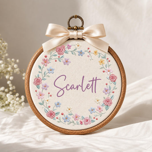 May include: A circular wooden embroidery hoop containing off-white fabric with a floral wreath design in pink, blue, and yellow thread. The name Scarlett is embroidered in purple script in the center. A cream satin bow sits at the top.