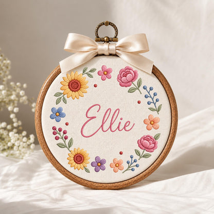 May include: A circular wooden embroidery hoop containing white fabric with the name Ellie embroidered in pink script. The name is surrounded by a colorful floral wreath featuring yellow sunflowers, pink peonies, blue flowers, and green leaves, topped with a cream satin bow.