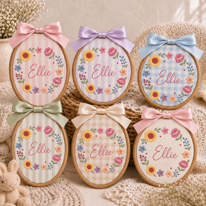 May include: Six oval wooden embroidery hoops featuring the name Ellie stitched in pink cursive script. Each hoop displays a floral wreath of sunflowers and small blossoms on various pastel striped or checkered fabrics, topped with a satin bow.