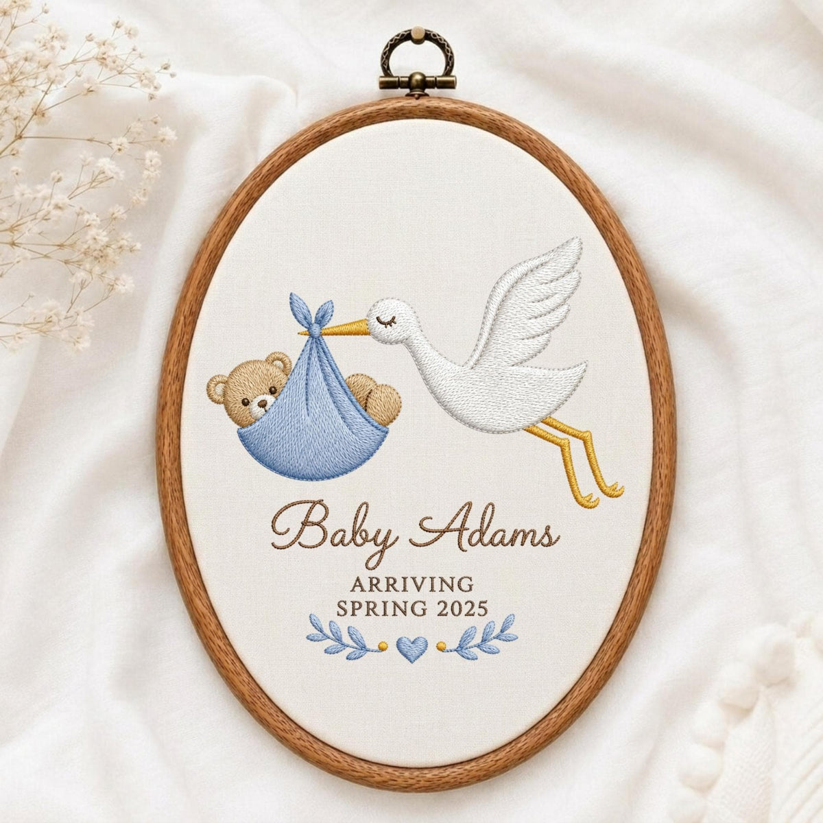 May include: An oval wooden embroidery hoop containing white fabric with a stitched design of a white stork carrying a brown teddy bear in a blue bundle. The text reads Baby Adams ARRIVING SPRING 2025, accented by blue leaves and a small heart.