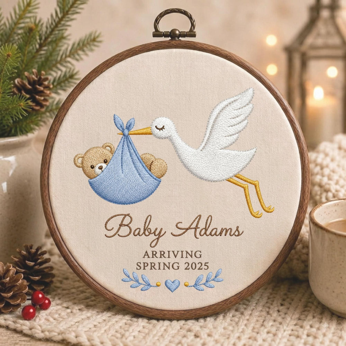 May include: A circular wooden embroidery hoop featuring a white fabric background with a stitched design of a white stork carrying a light blue bundle containing a brown teddy bear. The text reads Baby Adams ARRIVING SPRING 2025 with blue leaves.