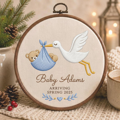 May include: A circular wooden embroidery hoop featuring a white fabric background with a stitched design of a white stork carrying a light blue bundle containing a brown teddy bear. The text reads Baby Adams ARRIVING SPRING 2025 with blue leaves.