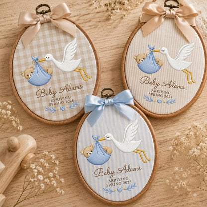 May include: Three oval wooden embroidery hoops featuring fabric backgrounds with beige gingham, beige stripes, and blue stripes. Each hoop displays an embroidered white stork carrying a blue bundle with a teddy bear, text reading Baby Adams ARRIVING SPRING 2025, and a satin bow.