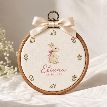 May include: A circular wooden embroidery hoop containing cream-colored fabric with an embroidered beige rabbit wearing a pink ribbon, surrounded by small floral sprigs. The name Eliana and the date 06.24.2027 are stitched below, topped with a cream satin bow.