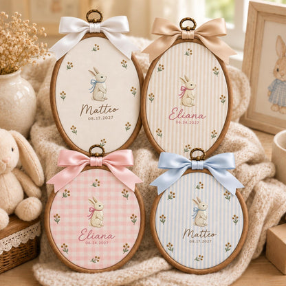 May include: Four oval wooden embroidery hoops featuring fabric inserts with printed bunny illustrations, floral accents, and personalized names and dates. Each hoop is topped with a decorative satin ribbon bow in white, beige, pink, or blue.