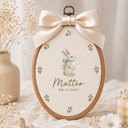 May include: An oval wooden embroidery hoop containing cream-colored fabric with a central embroidered rabbit wearing a blue ribbon, surrounded by small floral sprigs. The text reads Matteo and 08.17.2027, topped with a large beige satin bow.