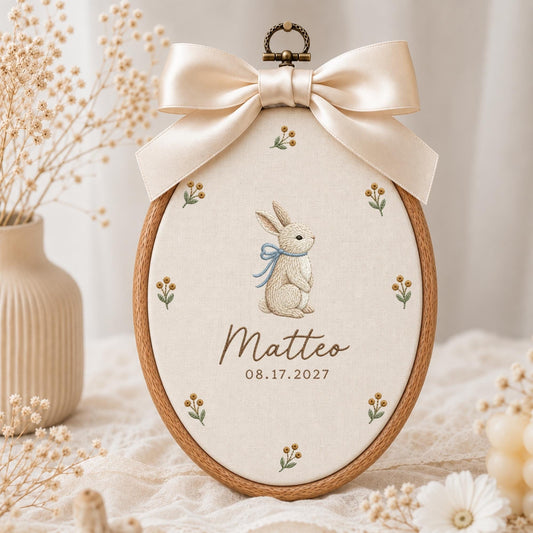 May include: An oval wooden embroidery hoop containing cream-colored fabric with a central embroidered rabbit wearing a blue ribbon, surrounded by small floral sprigs. The text reads Matteo and 08.17.2027, topped with a large beige satin bow.