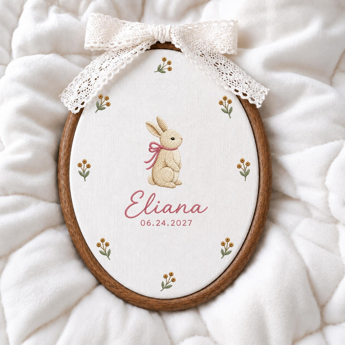 May include: An oval wooden embroidery hoop containing white fabric with a stitched beige rabbit wearing a pink ribbon, surrounded by small floral sprigs. The name Eliana and the date 06.24.2027 are embroidered in pink. A white lace bow is attached.