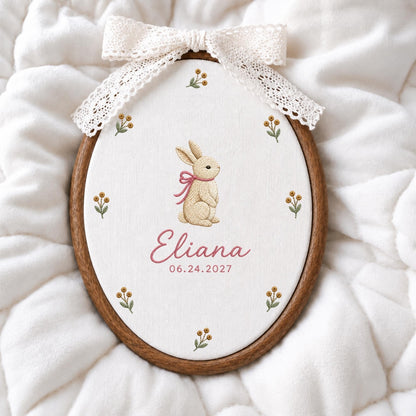 May include: An oval wooden embroidery hoop containing white fabric with a stitched beige rabbit wearing a pink ribbon, surrounded by small floral sprigs. The name Eliana and the date 06.24.2027 are embroidered in pink. A white lace bow is attached.