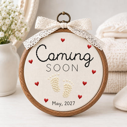 May include: A circular wooden embroidery hoop containing off-white fabric with black embroidered text reading Coming SOON and May, 2027. The design features small red embroidered hearts and cream-colored French knot baby footprints, topped with a lace bow.