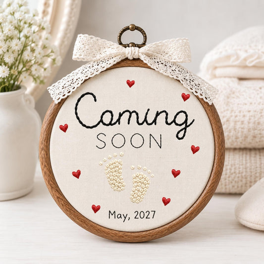 May include: A circular wooden embroidery hoop containing off-white fabric with black embroidered text reading Coming SOON and May, 2027. The design features small red embroidered hearts and cream-colored French knot baby footprints, topped with a lace bow.