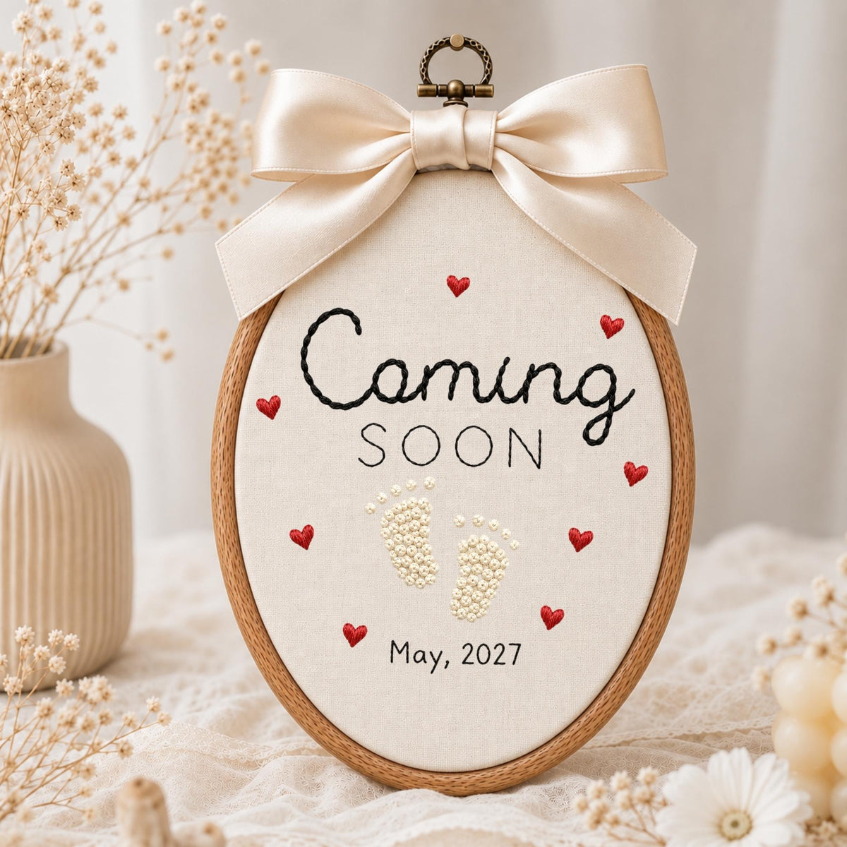 May include: An oval embroidery hoop featuring cream fabric with the black embroidered text Coming SOON, small red stitched hearts, white beaded baby footprints, and the date May, 2027. A large, smooth, champagne-colored satin bow sits at the top.