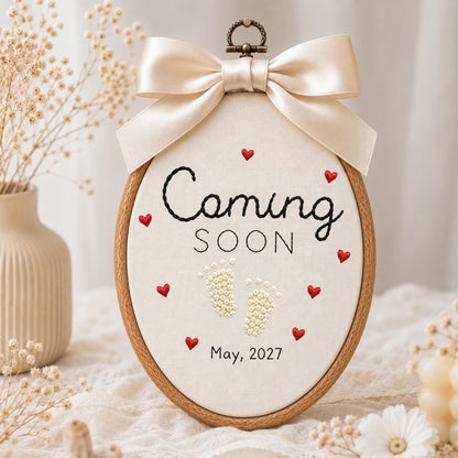 May include: An oval embroidery hoop featuring cream fabric with the black embroidered text Coming SOON, small red stitched hearts, white beaded baby footprints, and the date May, 2027. A large, smooth, champagne-colored satin bow sits at the top.