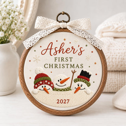 May include: A circular wooden embroidery hoop containing fabric with the embroidered text Asher's FIRST CHRISTMAS 2027. The design features three snowmen with orange noses, colorful hats, and scarves, topped with a white lace bow and a metal hanging ring.