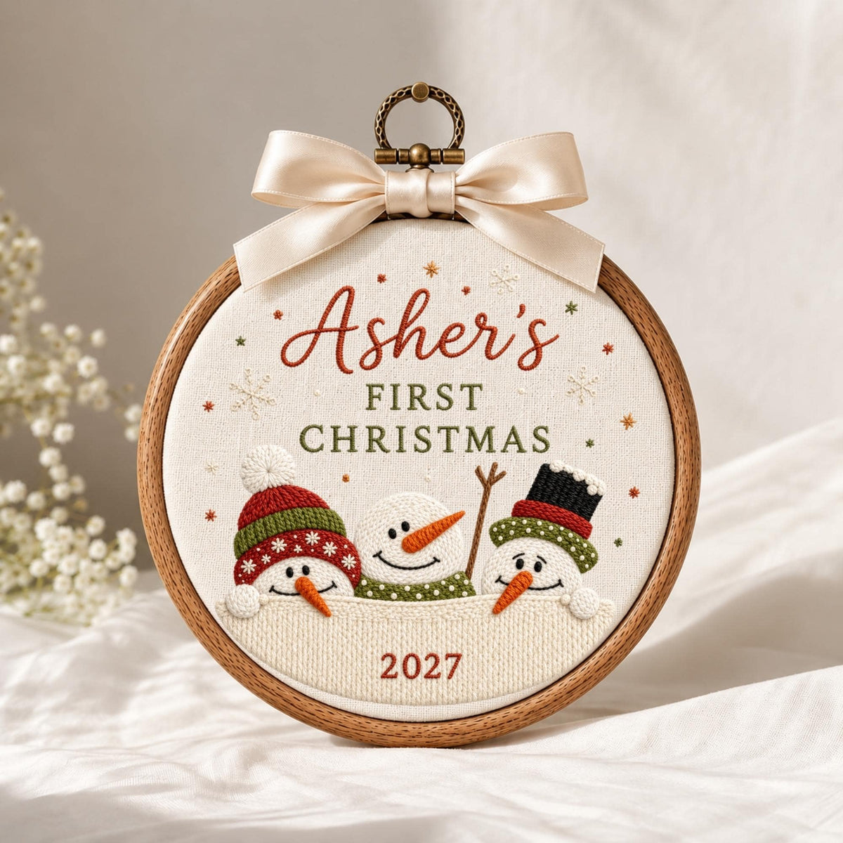 May include: A round wooden embroidery hoop containing white fabric with the embroidered text "Asher's FIRST CHRISTMAS 2027" and three snowmen wearing hats. A cream-colored satin ribbon bow is attached to the top metal hanging loop.