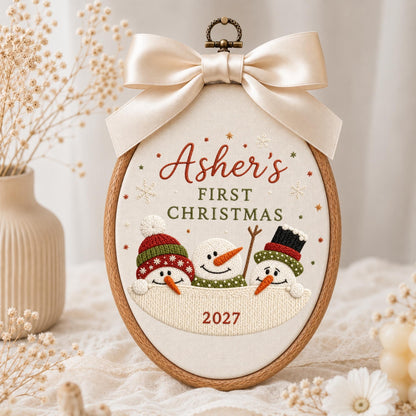 May include: An oval wooden embroidery hoop containing fabric with the embroidered text Asher's FIRST CHRISTMAS and 2027. Three snowmen with orange noses and colorful hats are stitched below the text. A large cream satin bow sits at the top.