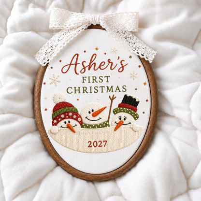 May include: An oval embroidery hoop featuring white fabric with the text Asher's FIRST CHRISTMAS and 2027. Three embroidered snowmen with orange noses and colorful hats are displayed. A white lace ribbon is tied at the top of the wooden frame.