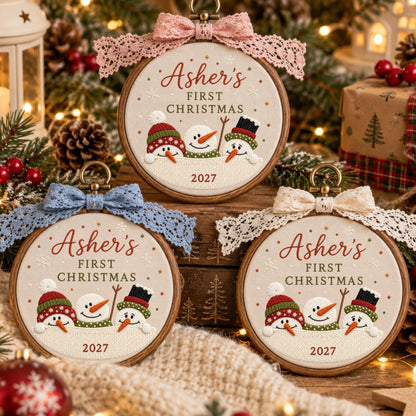 May include: Three circular wooden embroidery hoops featuring fabric with embroidered snowmen wearing colorful hats. The text reads Asher's FIRST CHRISTMAS 2027. Each hoop is topped with a decorative lace bow in pink, blue, or cream.