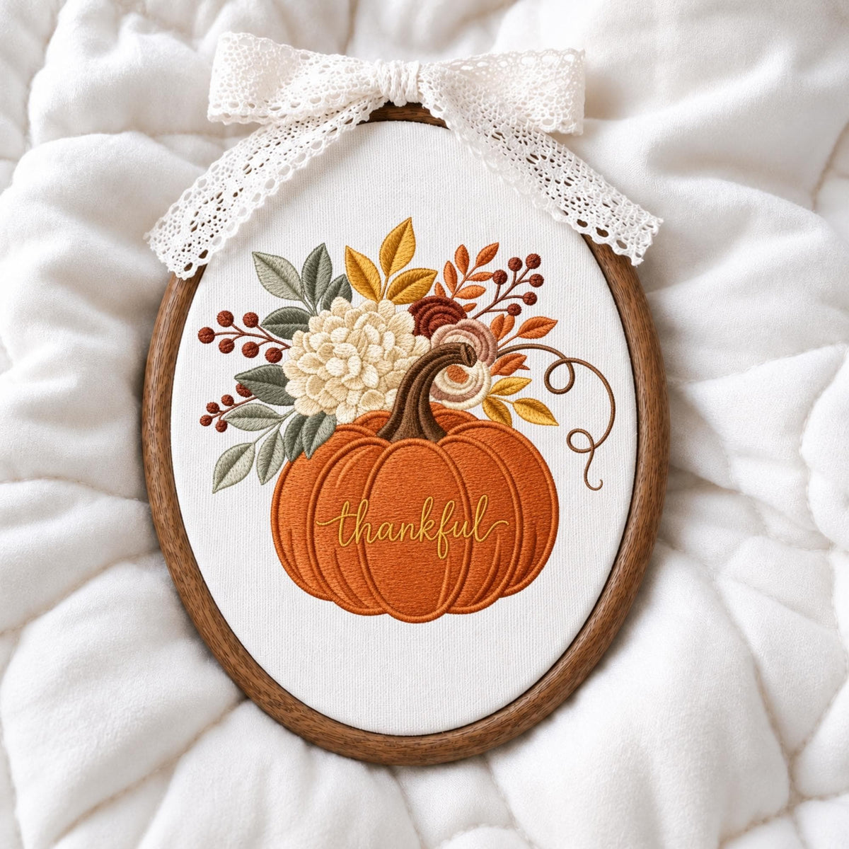 May include: An oval embroidery hoop containing white fabric with a stitched orange pumpkin featuring the word thankful in cursive script. The pumpkin is topped with embroidered cream flowers, green and yellow leaves, and brown berries, finished with a white lace bow.