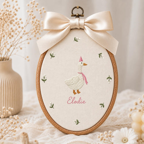 May include: An oval wooden embroidery hoop containing cream-colored fabric with a stitched white goose wearing a pink party hat and scarf. The name Elodie is embroidered in pink below the goose, surrounded by small floral sprigs and a satin bow.