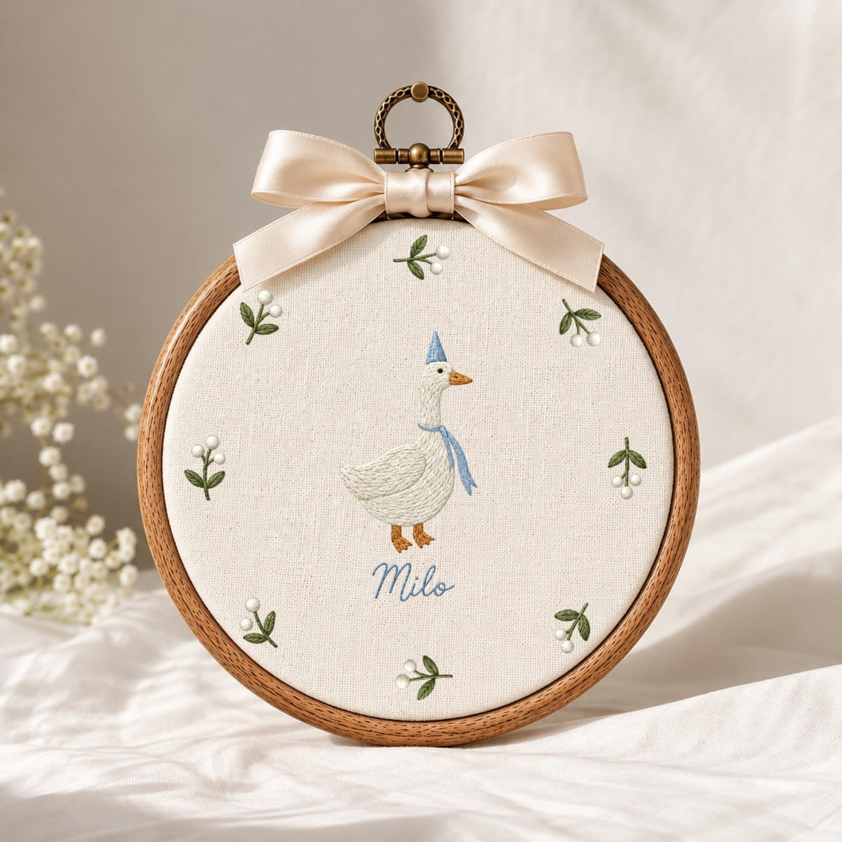 May include: A circular wooden embroidery hoop containing off-white fabric with a stitched white goose wearing a blue party hat and scarf. The name Milo is embroidered in blue below the goose, surrounded by small green sprigs with white berries.