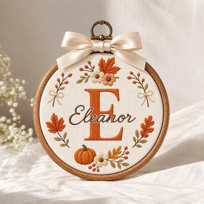 May include: A circular wooden embroidery hoop containing white fabric with an embroidered orange letter E and the name Eleanor in cursive script. The design features autumn leaves, flowers, and a pumpkin, topped with a cream satin ribbon bow.