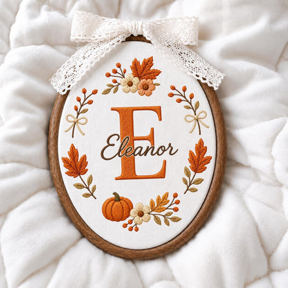 May include: An oval embroidery hoop containing white fabric with a large orange letter E and the name Eleanor embroidered in brown. The design features autumn leaves, berries, and a pumpkin, finished with a white lace bow at the top.