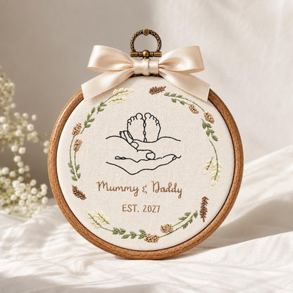 May include: A circular wooden embroidery hoop containing off-white fabric with a black line art design of baby feet held in hands. The text reads Mummy &amp; Daddy EST. 2027. A wreath of embroidered green leaves and brown wheat surrounds it, topped with a cream ribbon.