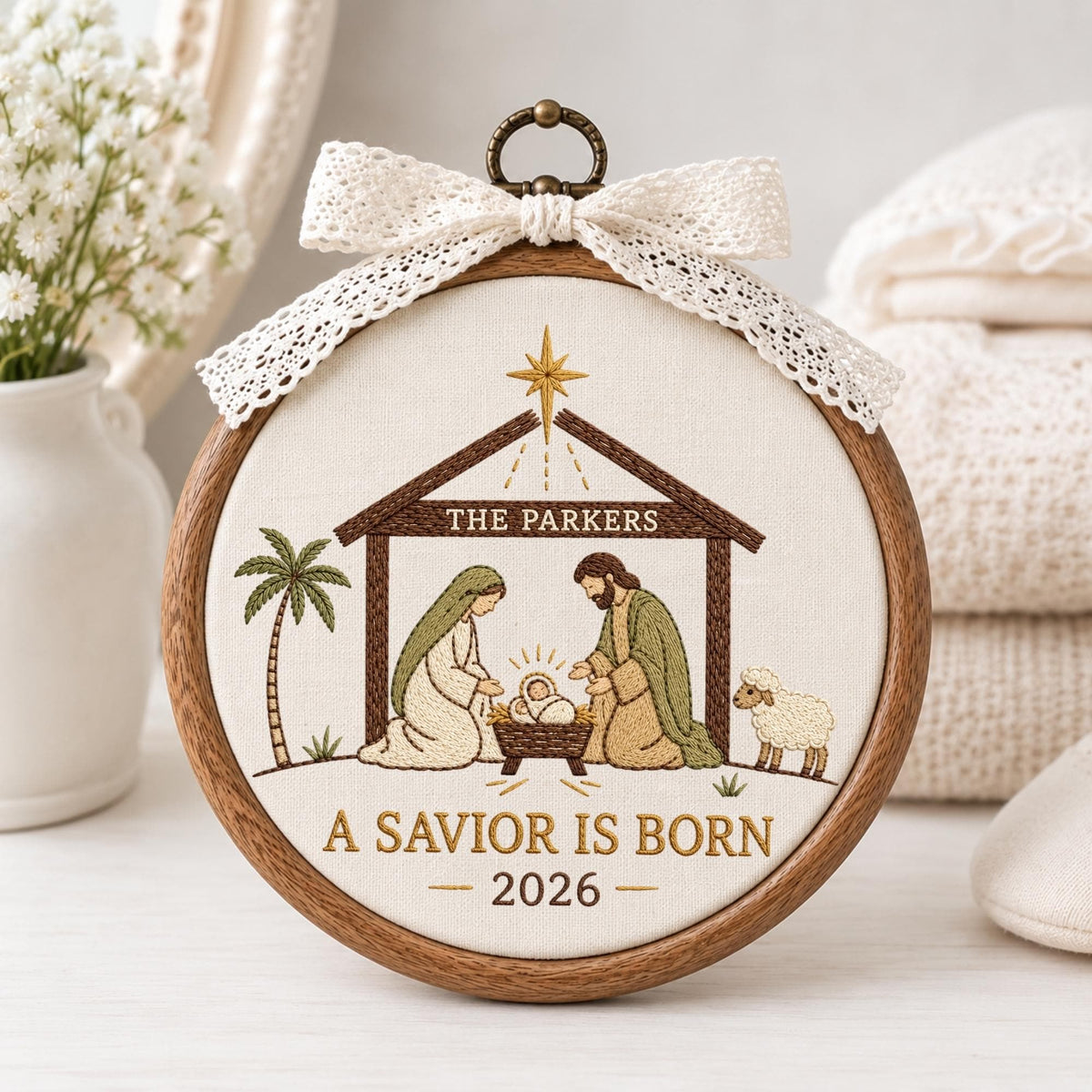 May include: A circular wooden embroidery hoop featuring a nativity scene stitched on white fabric. The design includes a stable, a palm tree, a sheep, and the text THE PARKERS, A SAVIOR IS BORN, and 2026. A white lace bow is attached to the top.