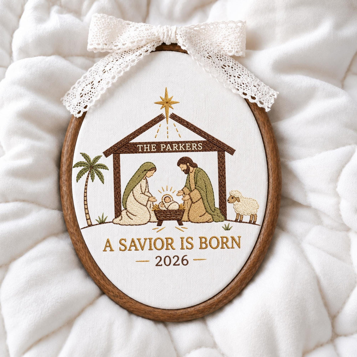 May include: An oval embroidery hoop featuring a nativity scene with a brown wooden frame and a white lace bow at the top. The embroidery depicts a stable, a palm tree, a sheep, and the text THE PARKERS, A SAVIOR IS BORN, and the year 2026.