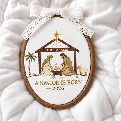 May include: An oval embroidery hoop featuring a nativity scene with a brown wooden frame and a white lace bow at the top. The embroidery depicts a stable, a palm tree, a sheep, and the text THE PARKERS, A SAVIOR IS BORN, and the year 2026.