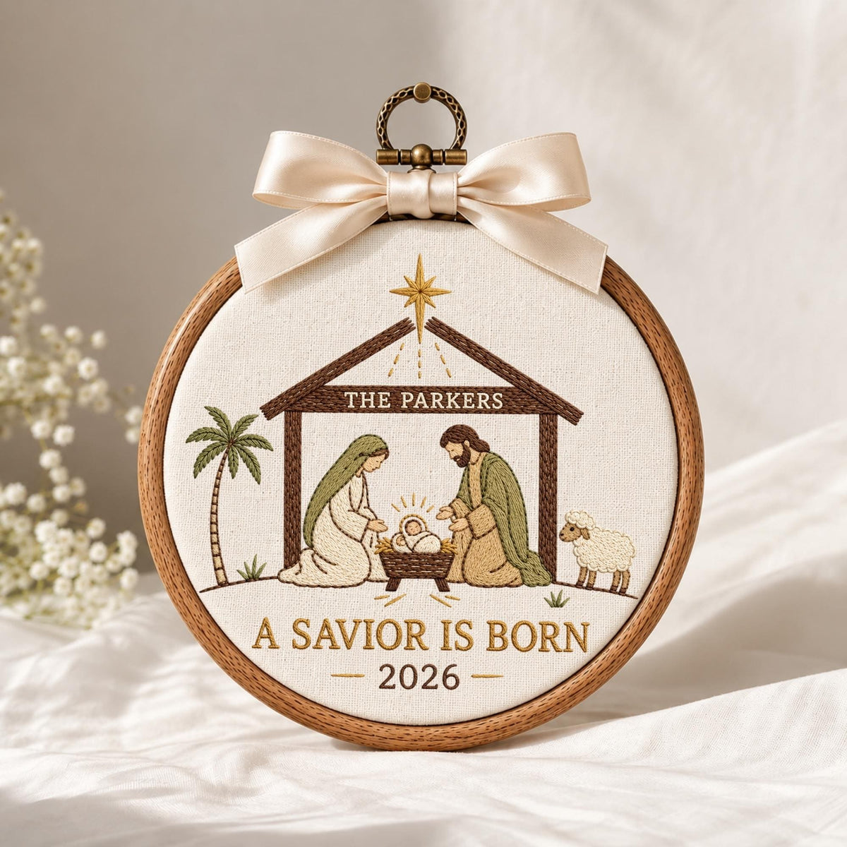 May include: A circular wooden embroidery hoop containing a fabric panel with a nativity scene. The design features a brown stable, a palm tree, a sheep, and figures in robes. Text reads THE PARKERS, A SAVIOR IS BORN, and 2026. A cream satin bow is attached to the top metal hanging loop.