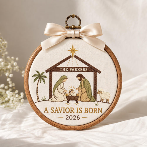 May include: A circular wooden embroidery hoop containing a fabric panel with a nativity scene. The design features a brown stable, a palm tree, a sheep, and figures in robes. Text reads THE PARKERS, A SAVIOR IS BORN, and 2026. A cream satin bow is attached to the top metal hanging loop.