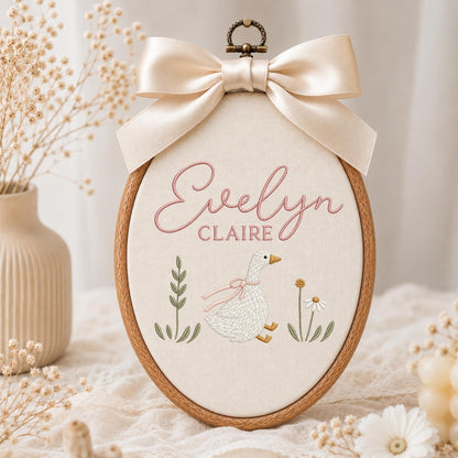 May include: An oval wooden embroidery hoop containing cream-colored fabric with the embroidered name Evelyn Claire in pink script. Below the name is a white goose with a pink ribbon, flanked by green botanical stems and a small white flower.