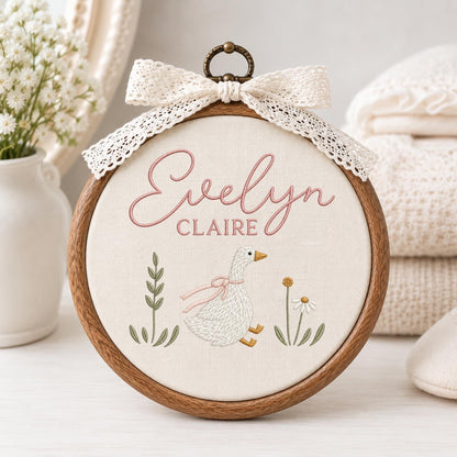 May include: A circular wooden embroidery hoop containing cream-colored fabric with the embroidered name Evelyn Claire in pink script and block letters. Below the name is a white goose with a pink ribbon, flanked by green foliage and a daisy.