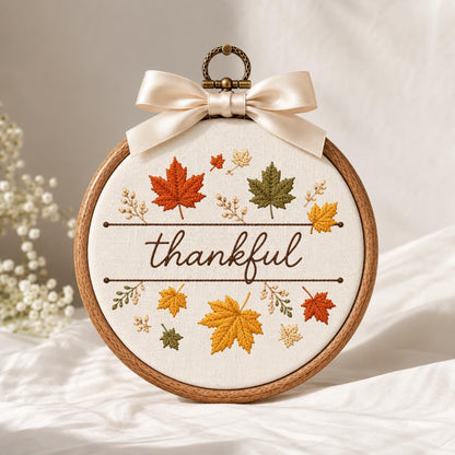 May include: A circular wooden embroidery hoop containing white fabric with the embroidered word thankful in brown script. The design features colorful maple leaves in shades of orange, yellow, and green, accented by a cream-colored satin ribbon bow.