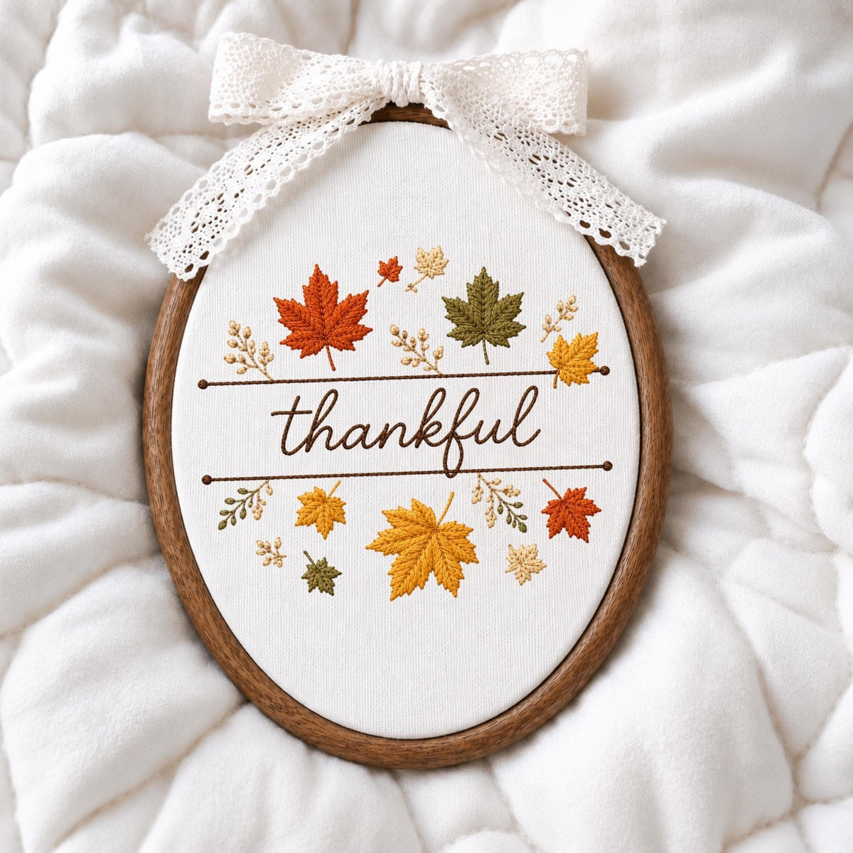 May include: An oval embroidery hoop containing white fabric with the word thankful stitched in brown thread. The design features embroidered maple leaves in shades of orange, yellow, and green, accented by a white lace bow attached to the top.