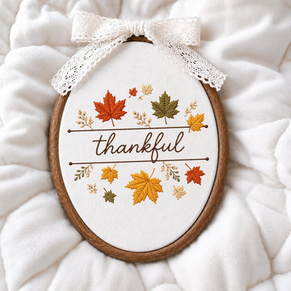 May include: An oval embroidery hoop containing white fabric with the word thankful stitched in brown thread. The design features embroidered maple leaves in shades of orange, yellow, and green, accented by a white lace bow attached to the top.