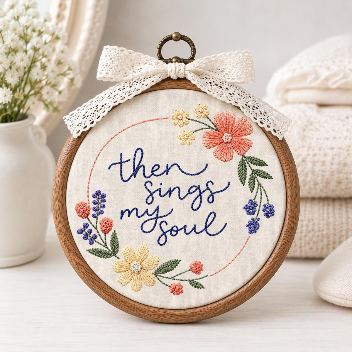 May include: A circular wooden embroidery hoop containing off-white fabric with the hand-stitched dark blue text "then sings my soul" surrounded by a floral wreath of pink, yellow, and purple flowers, topped with a cream-colored lace bow.