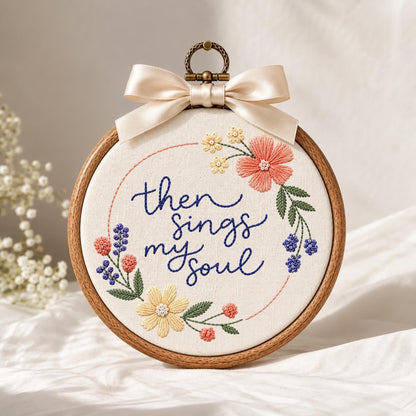 May include: A circular wooden embroidery hoop containing white fabric with the hand-stitched dark blue text "then sings my soul" surrounded by a floral wreath of pink, yellow, and blue flowers with green leaves, topped with a cream satin bow.