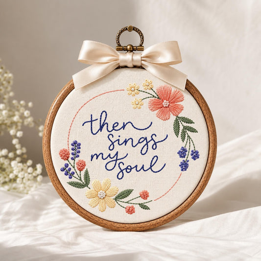 May include: A circular wooden embroidery hoop containing white fabric with the hand-stitched dark blue text "then sings my soul" surrounded by a floral wreath of pink, yellow, and blue flowers with green leaves, topped with a cream satin bow.